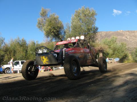 1990 Ford Bronco - red tube-frame off-road race buggy with a yellow lion emblem gets airborne off a dirt rise under a clear blue sky.