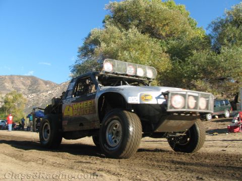 1990 Ford Bronco - off-road race truck lettered '4 Wheel Parts' lifts a front wheel as it powers along a dirt course past trees and pits.