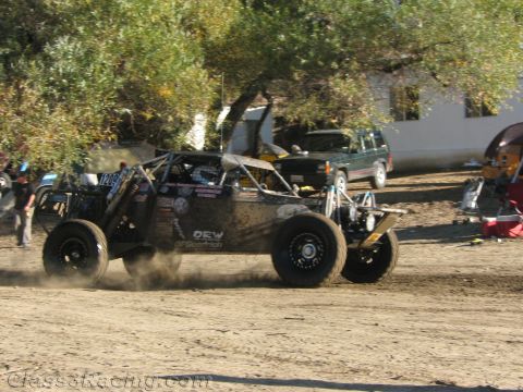 1990 Ford Bronco - black single-seat off-road race buggy throws up dust on a dirt track with spectators and a building in the background.