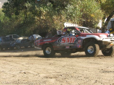 1990 Ford Bronco - white and red prerunner-style race truck marked SMI churns across packed dirt past parked vehicles and trees.
