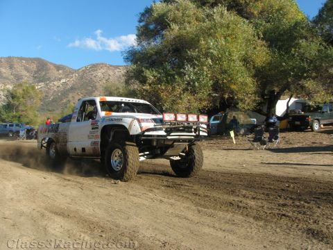 1990 Ford Bronco - white sponsor-covered race truck catches air over a dirt rise with mountains and trees behind it.