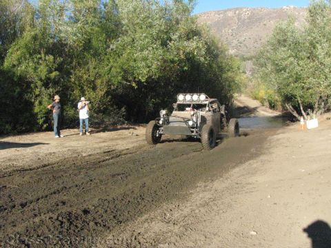 1990 Ford Bronco - light-bar-equipped off-road buggy heads toward a muddy water crossing while two people photograph from the bank.