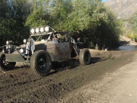 1990 Ford Bronco - tube-frame off-road buggy with a roof light bar drives along a muddy creekside trail beneath green trees.
