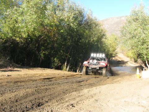 1990 Ford Bronco - red race truck with a roof light bar splashes through a shallow stream on a sunlit tree-lined dirt trail.