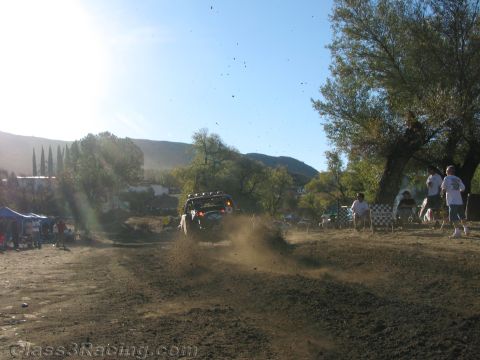 1990 Ford Bronco - backlit off-road vehicle throws up a cloud of dust heading toward spectators in a sunlit dirt clearing.