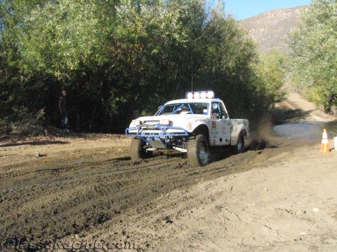 1990 Ford Bronco - white sponsor-marked race truck sprays mud as it drives a dirt trail past an orange traffic cone.