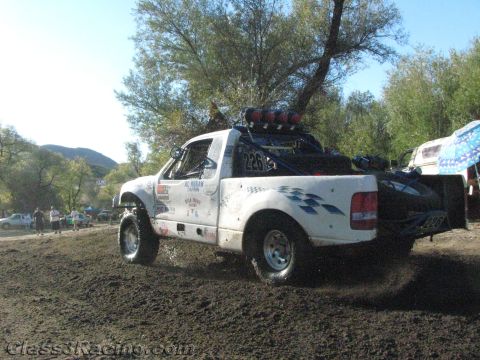 1990 Ford Bronco - Rear three-quarter view of white race truck number 226 driving across churned dirt with a roof light bar.
