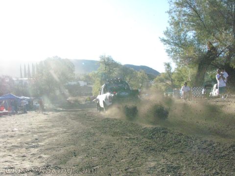1990 Ford Bronco - backlit off-road truck sprays dirt across a hazy sunlit clearing as spectators watch near tents and trees.