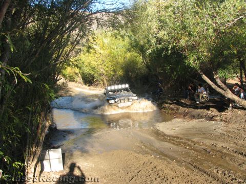 1990 Ford Bronco - light-bar-equipped off-road buggy throws up spray crossing a tree-shaded stream while spectators sit on the bank.