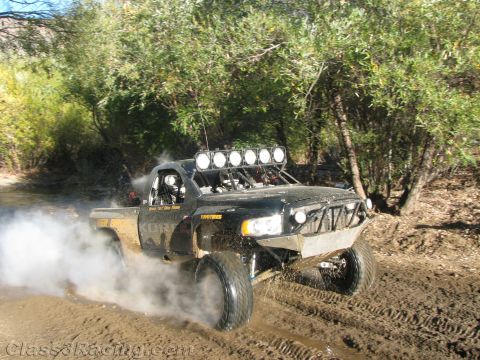 1990 Ford Bronco - black race truck with a six-light roof bar sprays water and mud while powering along a brushy dirt trail.