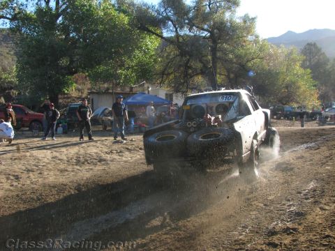 1990 Ford Bronco - White race truck number 910 sprays mud through a churned pit area as spectators watch under shade trees.