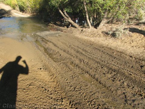1990 Ford Bronco - muddy trail scored with tire tracks runs beside a stream, the camera's shadow cast across the foreground.