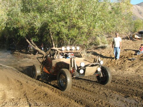 1990 Ford Bronco - open tube-frame sand-rail buggy with a light bar climbs a muddy berm as bystanders watch beside trees.