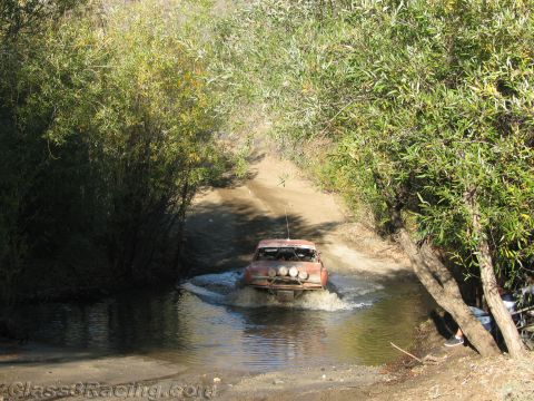 1990 Ford Bronco - orange off-road vehicle splashes through a shaded stream crossing on a narrow tree-lined dirt trail.