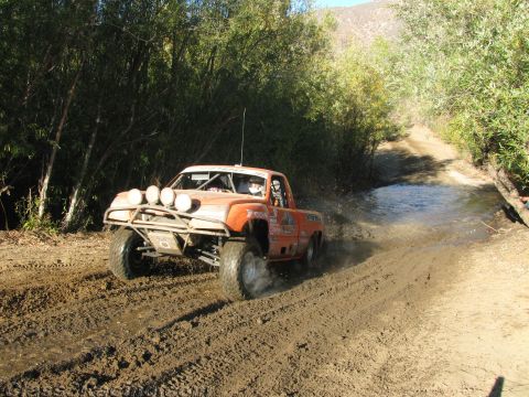 1990 Ford Bronco - orange race truck with a roof light bar sprays mud powering along a brushy dirt trail beside a stream.