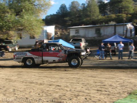1990 Ford Bronco - white and red race truck drives a dirt course past spectators, tents, and parked trucks near a building.