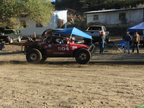 1990 Ford Bronco - red off-road race buggy numbered 504 runs a dirt course past a blue canopy and watching spectators.