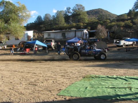 1990 Ford Bronco - blue off-road race buggy numbered 518 drives a dirt course past pit canopies with green hills behind.