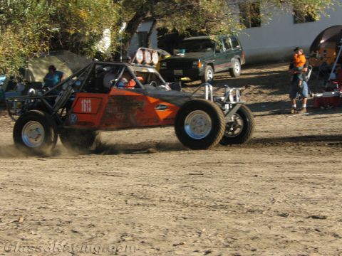1990 Ford Bronco - orange and black off-road buggy numbered 1513 slides through dirt past a parked SUV and spectators.