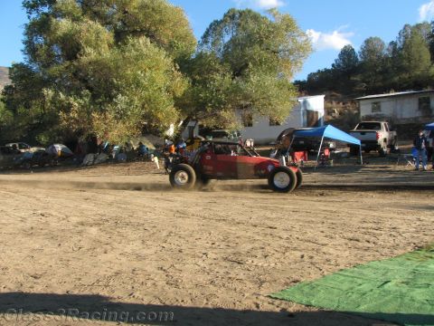 1990 Ford Bronco - red off-road race buggy kicks up dust crossing a dirt pit area past canopies, parked trucks, and trees.