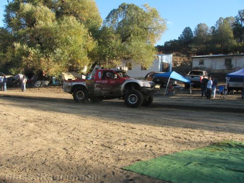 1990 Ford Bronco - red and white race truck drives a dirt course beside blue pit canopies and parked vehicles under trees.