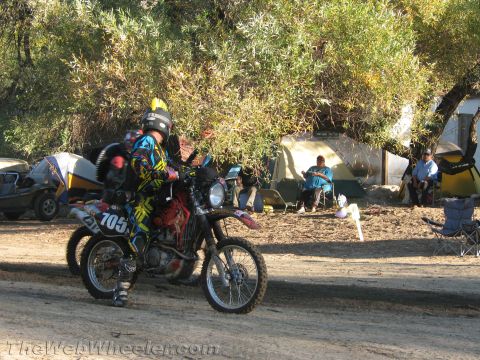 1990 Ford Bronco - motorcyclist in colorful gear rides dirt bike number 105 along a dusty desert trail beside a campsite with tents and onlookers.