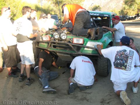 1990 Ford Bronco - pit crew swarms a green tube-frame off-road race truck on a dirt lot, repairing the front end with one mechanic on top.