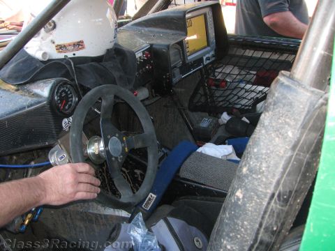 1990 Ford Bronco - stripped interior of an off-road race car shows a suede steering wheel, a mounted GPS, gauges and a roll cage with a hand on the wheel.