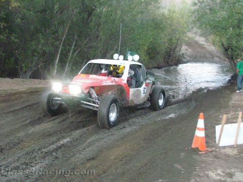 1990 Ford Bronco - red and white off-road race buggy with roof-mounted lights powers through a muddy creek crossing beside an orange traffic cone.