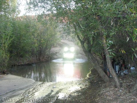 1990 Ford Bronco - off-road vehicle's headlights glare as it splashes through a calm creek framed by willow trees at dusk.