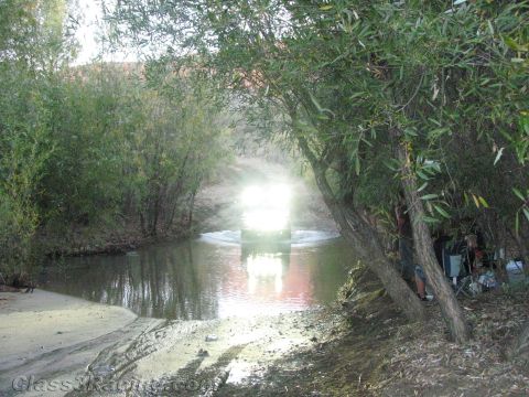 1990 Ford Bronco - Bright headlights of an off-road vehicle reflect off the water as it crosses a tree-lined creek in low evening light.