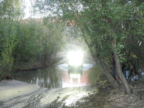 1990 Ford Bronco - off-road vehicle pushes through a shallow creek, its headlights flaring brightly against the dim wooded crossing.