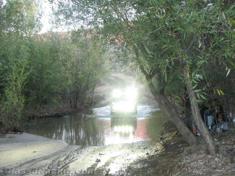 1990 Ford Bronco - Headlights blaze from an off-road vehicle as it fords a shallow creek bordered by willow trees at dusk.