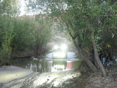 1990 Ford Bronco - off-road vehicle splashes across a shallow creek with headlights glowing, framed by overhanging willow branches.