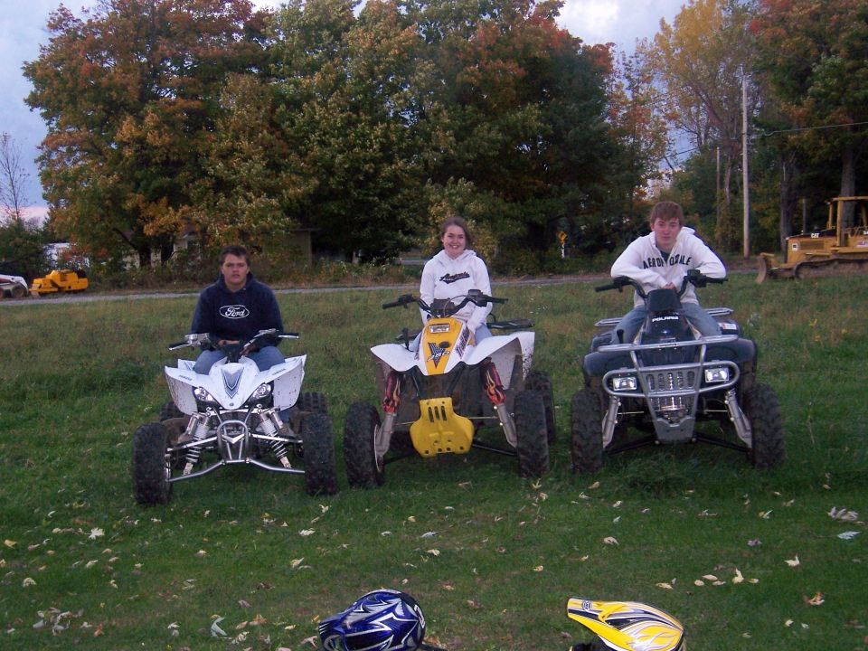 Three people seated on sport ATV quads on grass with autumn trees