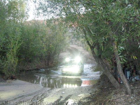 1990 Ford Bronco - off-road vehicle with several bright lights churns up muddy water as it exits a creek crossing among willow trees.