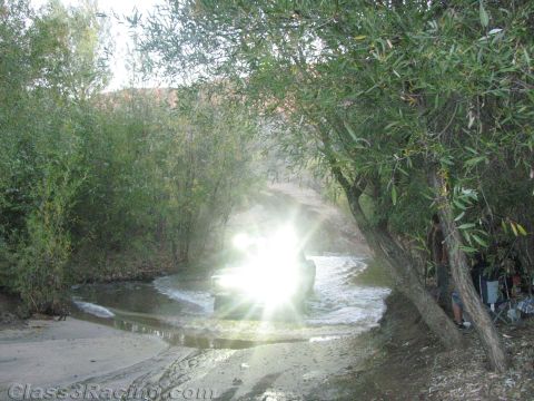 1990 Ford Bronco - off-road vehicle's lights create a starburst flare as it crosses a shallow creek bordered by willows at dusk.