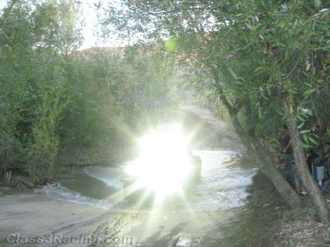 1990 Ford Bronco - intense starburst of light from an off-road vehicle's headlights dominates a dusk creek crossing among willow trees.