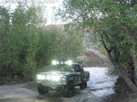 1990 Ford Bronco - black classic Ford pickup race truck on knobby tires crosses a shallow creek with roof and bumper lights blazing.