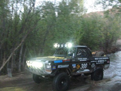 1990 Ford Bronco - black number 300 Ford off-road race truck with a roof light bar splashes through a creek, sponsor decals visible on its body.