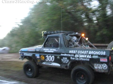 1990 Ford Bronco - black number 300 West Coast Broncos off-road race truck, carrying a spare tire in the bed, races down a wooded dirt road.