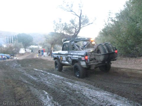 1990 Ford Bronco - black number 300 off-road race truck rounds a muddy rutted trail past a hillside spectator camp in dim evening light.