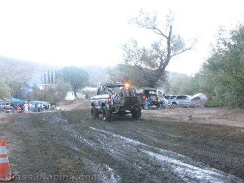 1990 Ford Bronco - off-road race truck heads down a deeply rutted muddy road past spectator tents and parked vehicles in fading dusk light.