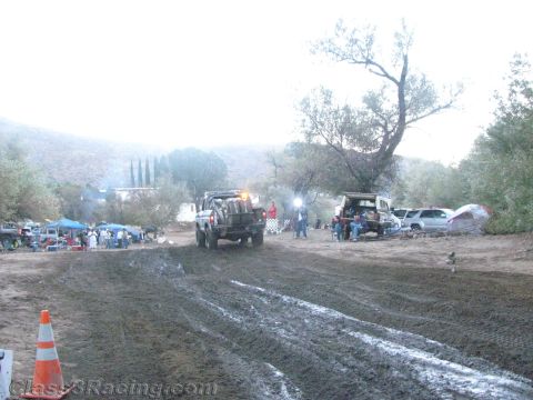 1990 Ford Bronco - pickup race truck drives along a muddy rutted course flanked by spectators, tents and parked cars under a misty dusk sky.
