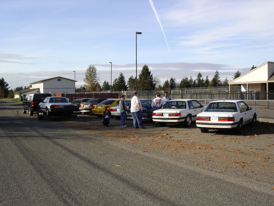 Several white Ford Mustangs parked in a lot, some people standing around them