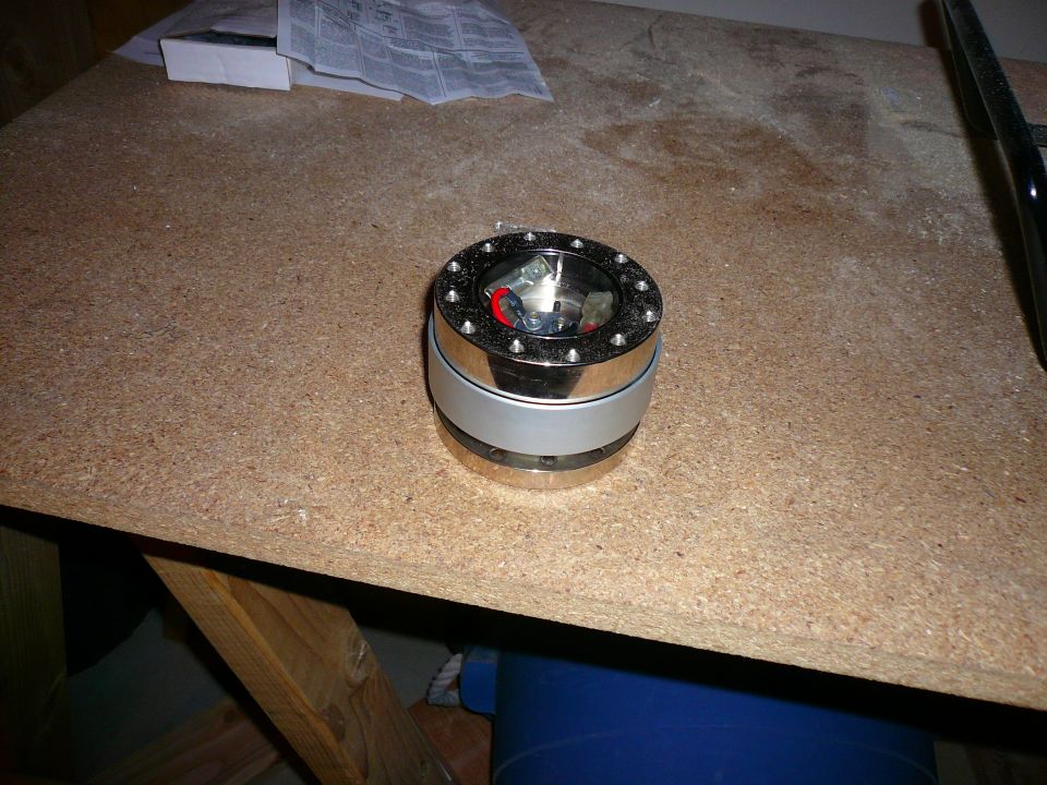 Chrome and silver steering wheel hub adapter with visible bolts and red accents, resting on a particle board surface