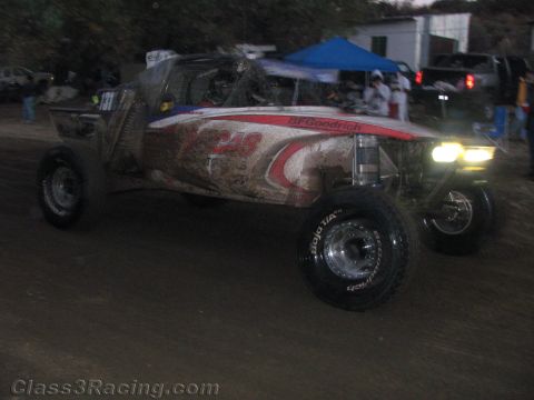 1990 Ford Bronco - mud-spattered white and red off-road race truck with a long-travel suspension races past a blue canopy pit area at dusk.