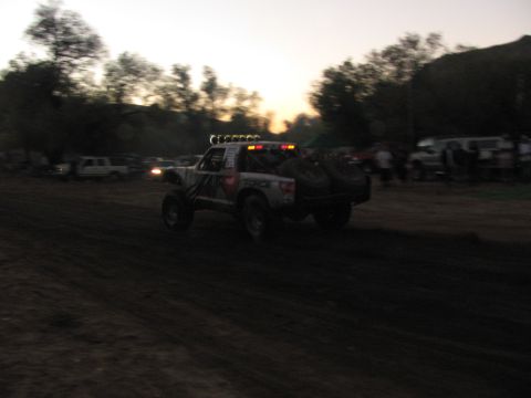 1990 Ford Bronco - off-road race truck with an illuminated roof light bar speeds across a dirt course against a glowing sunset sky.