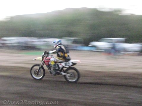 1990 Ford Bronco - motion-blurred rider races dirt bike number 172 across a dirt course with parked vehicles lining the background.