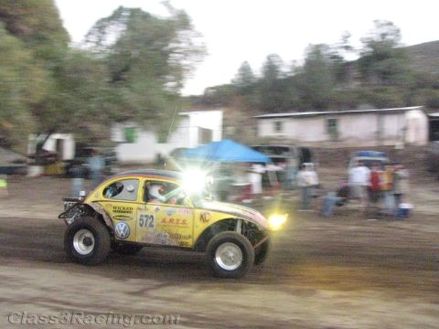 1990 Ford Bronco - yellow number 572 Volkswagen-based Baja Bug with KC lights races on a dirt course past a building and onlookers.